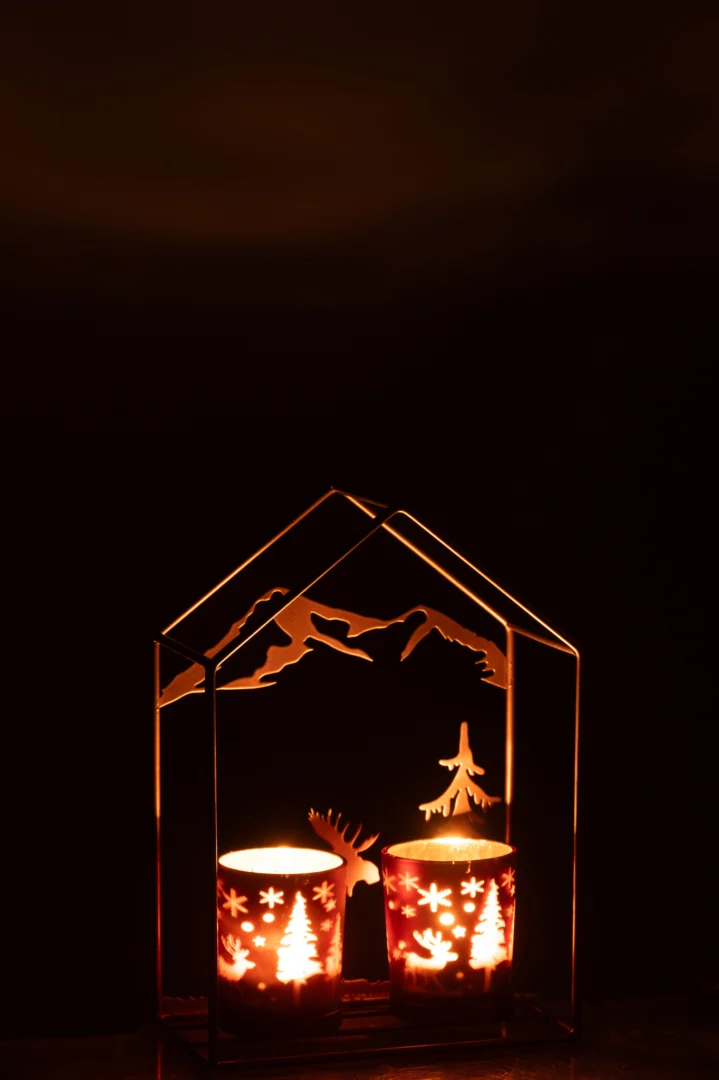 De Theelichthouder Huis IJzer Roestbruin/Bruin 2 Glazen is een huisvormige kandelaar met boom- en rendieruitsnijdingen, die zachte feestelijke schaduwen werpen op een zwarte achtergrond voor een warme gloed in het donker.