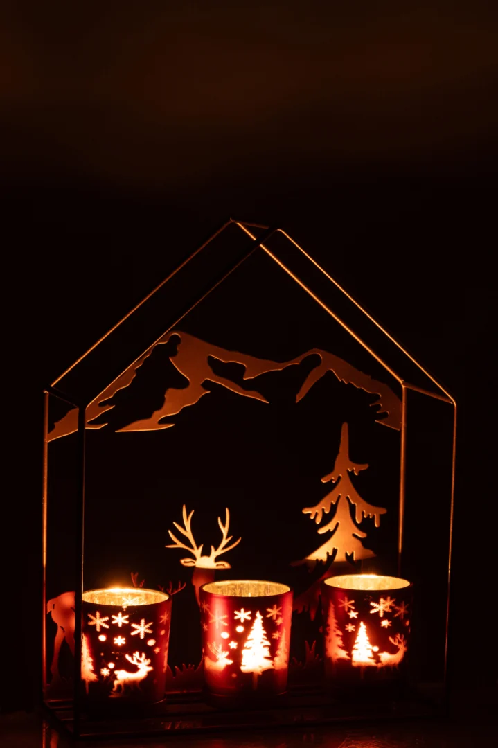 De Theelichthouder Huis IJzer Roestbruin/Bruin 3 Glazen heeft drie kaarsen in bruine glazen houders in een roestbruin metalen huis, met berg-, herten- en boomsilhouetten die een warme gloed in het donker werpen.