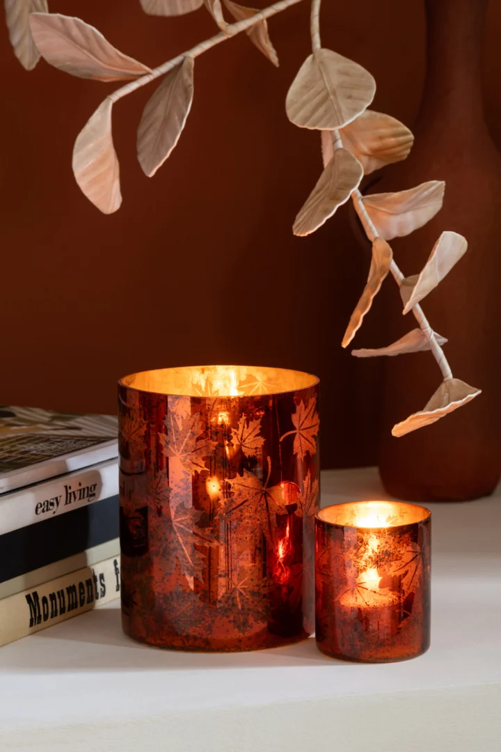 Twee brandende Kaarsenhouder Glas Roestbruin Klein kaarsen met bladmotieven staan op een wit oppervlak naast gestapelde boeken en decoratieve beige bladeren en creëren een warme, herfstige sfeer.