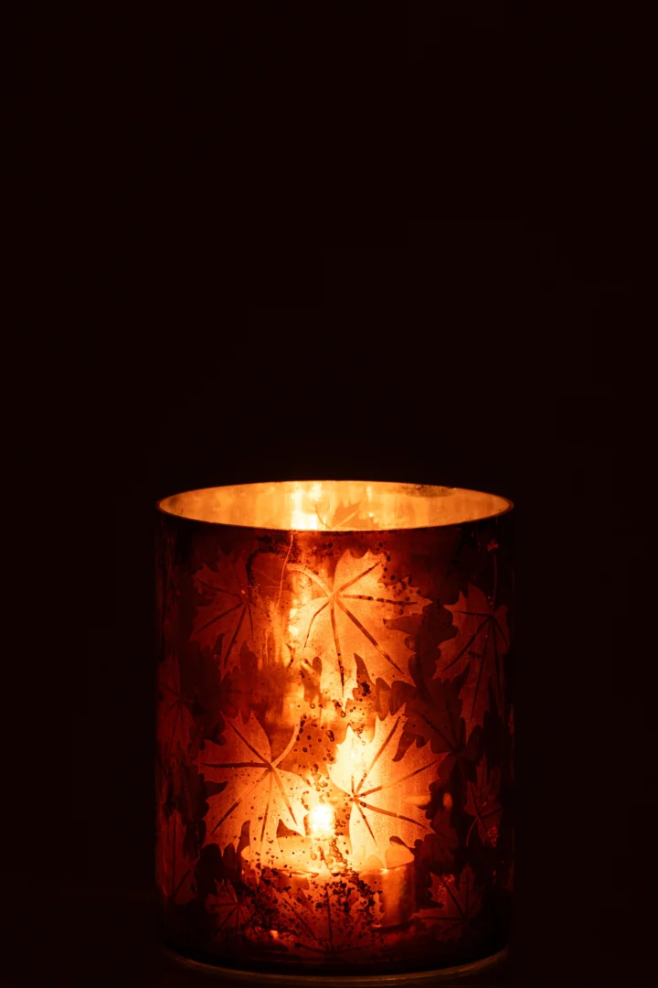 De Kaarsenhouder Glas Roestbruin Groot gloeit warm met een brandende kaars erin. De glazen houder met bladmotieven werpt een zacht oranje licht tegen een donkere achtergrond.