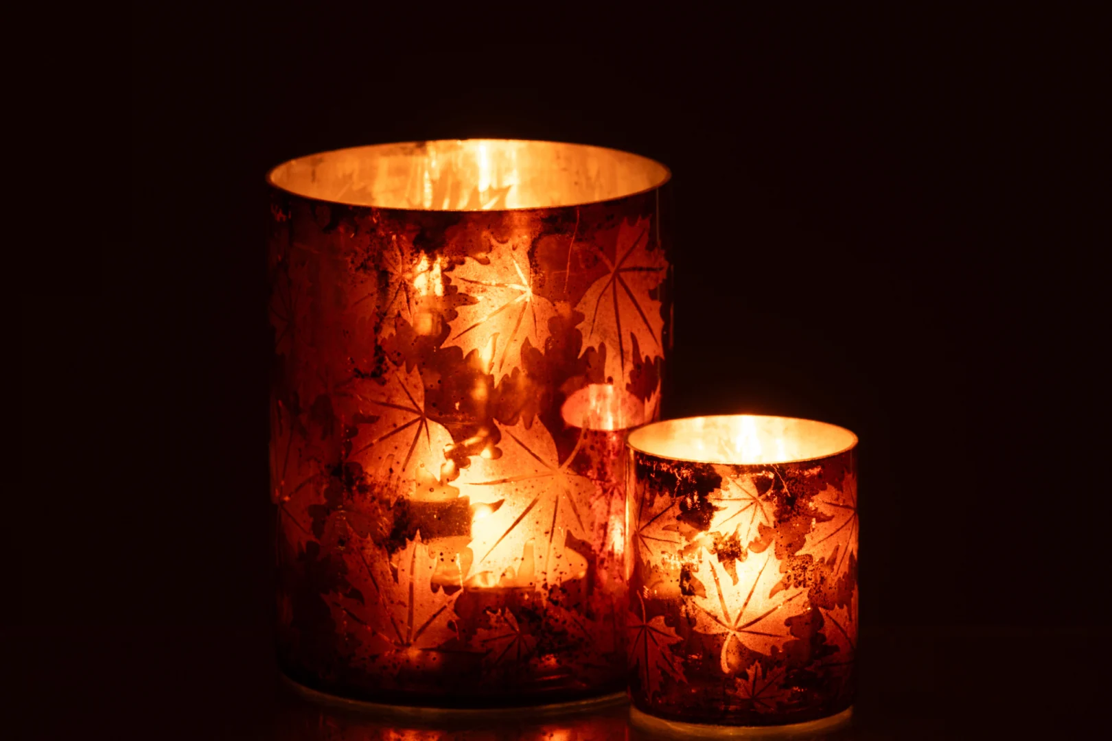 De Kaarsenhouder Glas Roestbruin Groot is een gloeiende glazen kandelaar met een herfstbladmotief, die een warm oranje licht uitstraalt. Links afgebeeld tegen een donkere achtergrond naast een kleinere bijpassende houder.