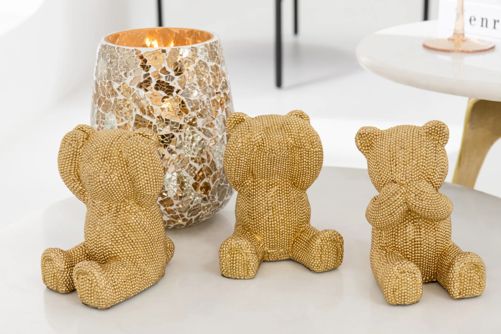 Drie gouden kralen teddybeer figuurtjes in het 'see no evil, hear no evil, speak no evil' motief zitten naast een brandende Kaarsenhouder Gebarsten Glas Goudkleurig kandelaar op een wit oppervlak.