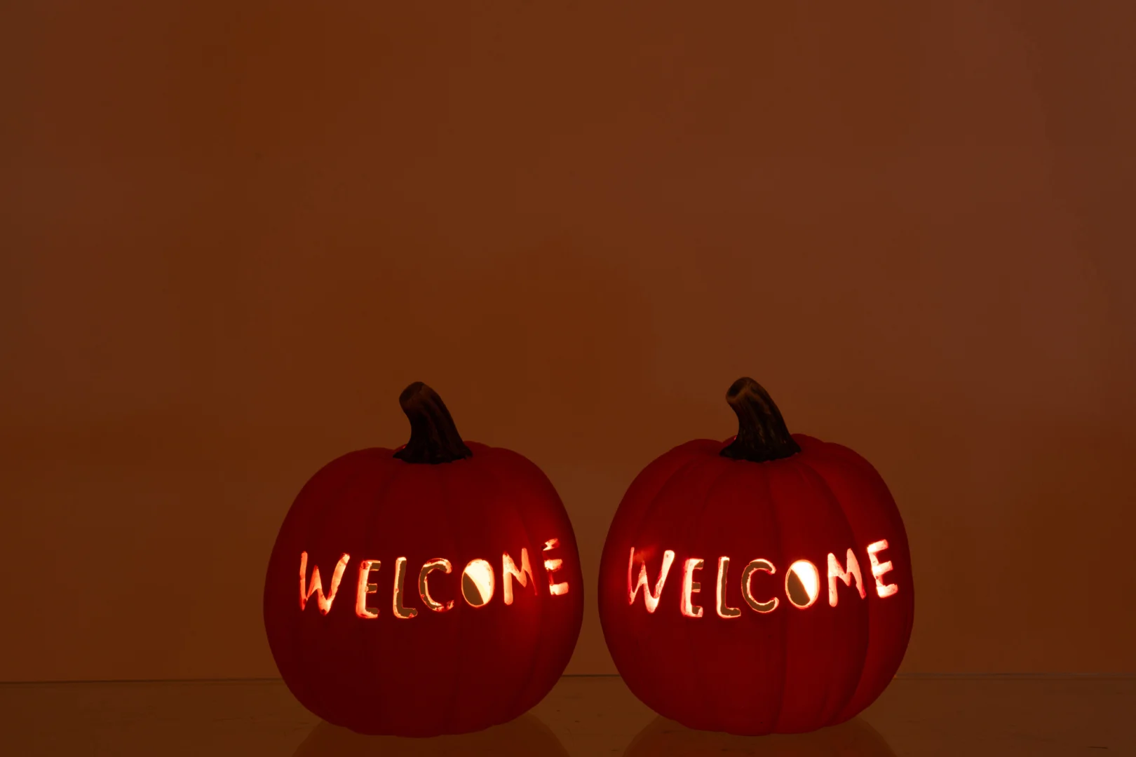 Set van 2 grote oranje kaarsenhouders in pompoenvorm, met het woord 'WELKOM' uitgesneden, die warm oplichten tegen een effen bruine achtergrond.