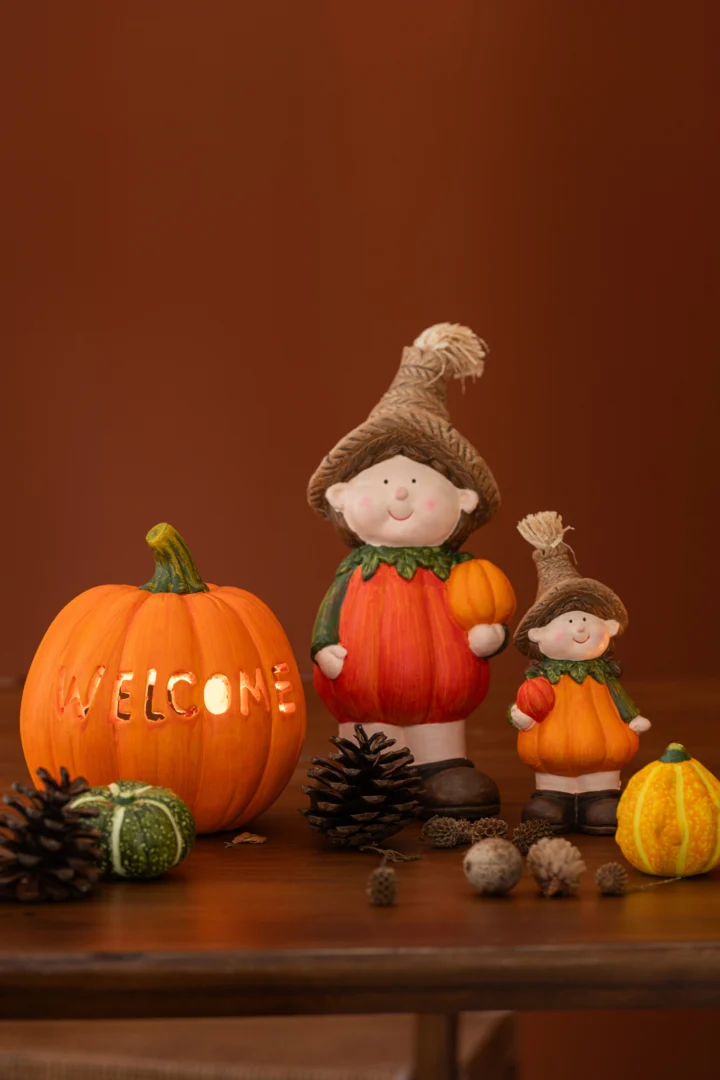 De Kaarsenhouder Pompoen Klei Welkom Oranje Donker Oranje Groot Set van 2 heeft twee pompoenkandelaars, een grote en een kleine, naast een bord met "WELKOM", met dennenappels, eikels en kalebassen op een houten ondergrond.