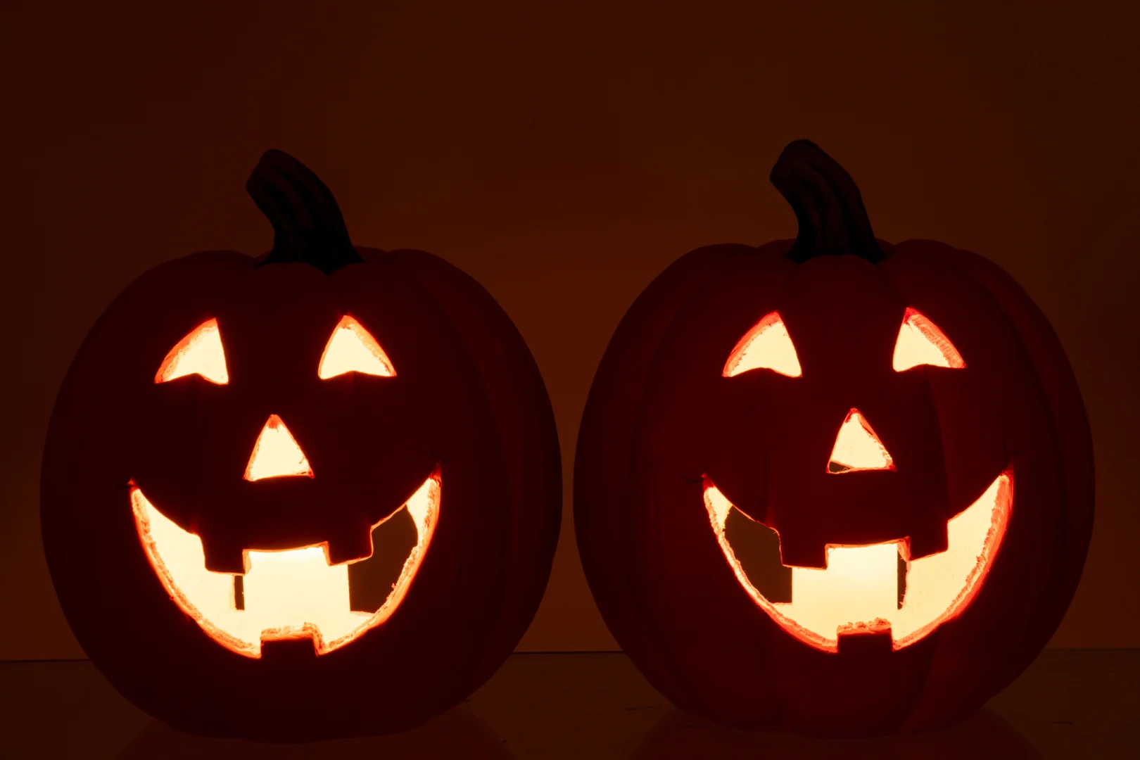 De Pompoen Halloween Magnesium Oranje/Donker Oranje Set van 2 heeft twee klassieke jack-o'-lantaarns met driehoekige ogen, neuzen en tandige glimlachen, gloeiend naast elkaar tegen een donkere achtergrond.
