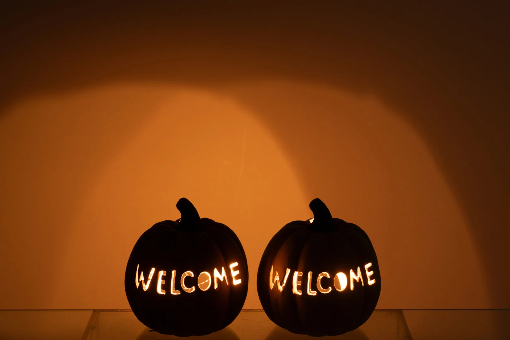 De Kaarsenhouder Pompoen Klei Welkom Karamel Groot heeft twee uitgesneden pompoenen die van binnenuit verlicht zijn en waarop in hoofdletters "WELKOM" staat, zij aan zij tegen een zacht verlichte oranje achtergrond.