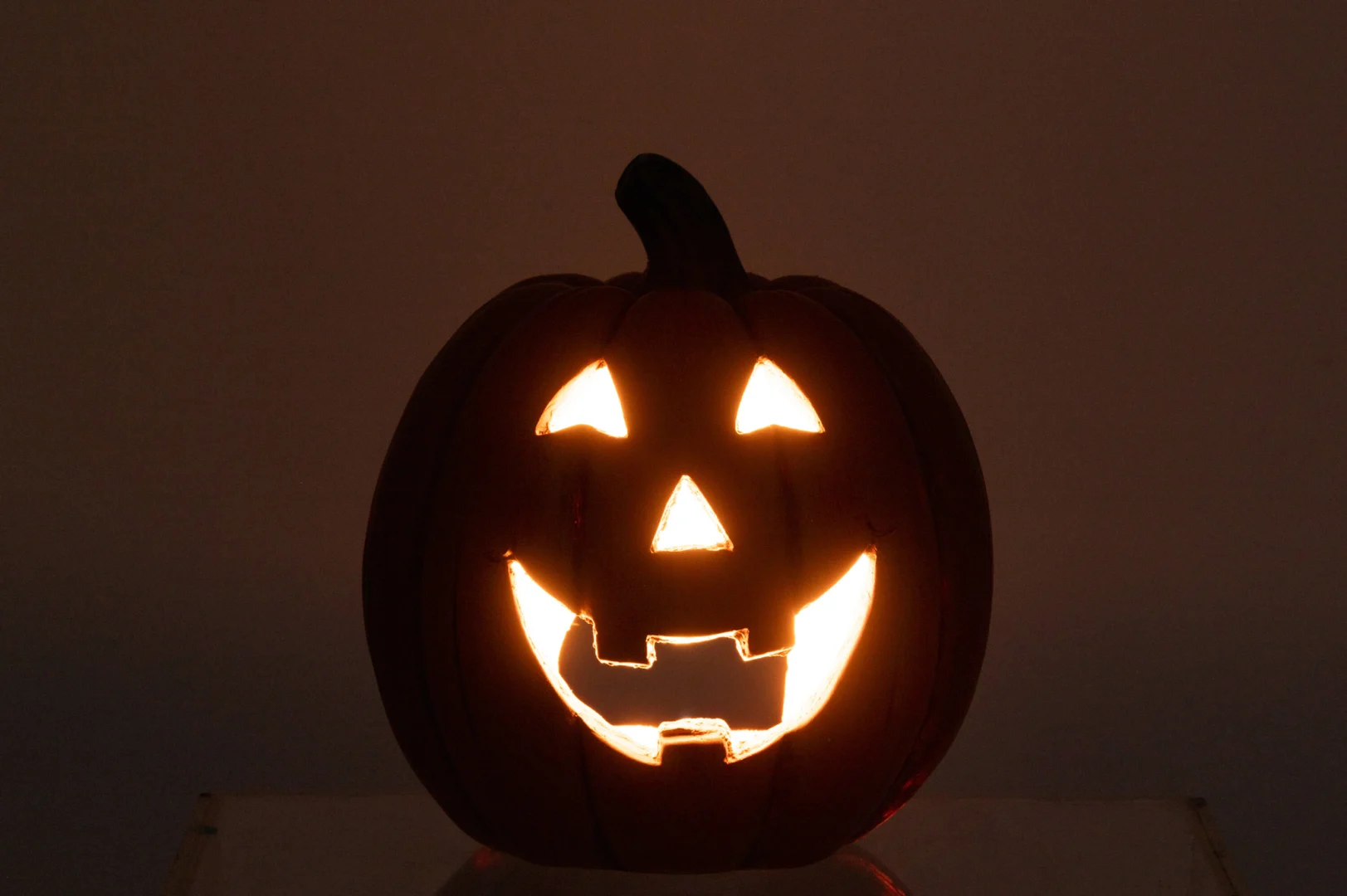 De Pompoen Halloween Klei Karamel heeft een klassiek jack-o'-lantern gezicht met driehoekige ogen, een driehoekige neus en een brede getande glimlach, die warm oplicht tegen een effen donkere achtergrond.