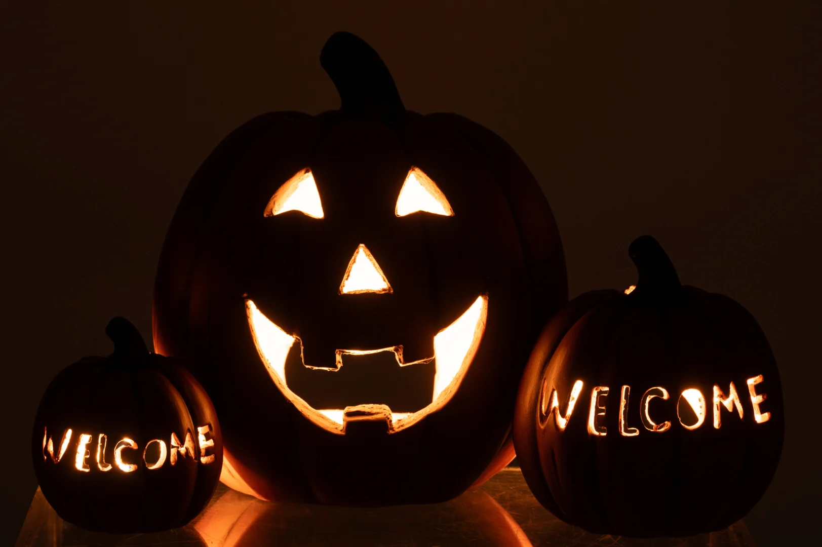 Drie "Pompoen Halloween Klei Karamel" jack-o'-lantaarns gloeien op een donkere achtergrond: de middelste pompoen heeft een klassiek uitgesneden gezicht, terwijl de kleinere pompoenen aan de zijkant zijn uitgesneden met "WELKOM".