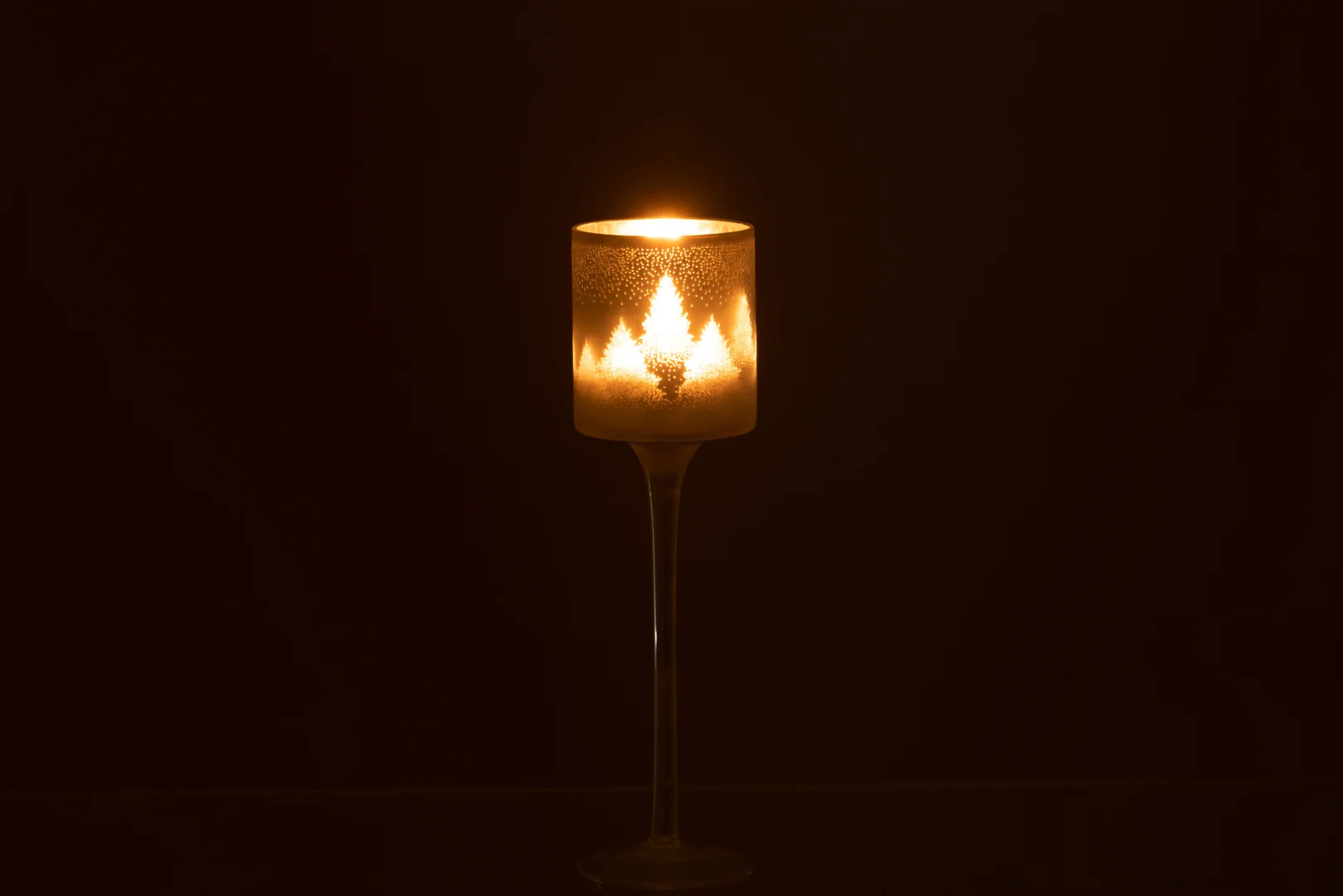 De Theelichthouder Denneboom Glas Grijs Zilver Klein houdt een enkele brandende kaars op een slanke steel, die een warme gloed en zachte schaduwen werpt tegen een donkere achtergrond.