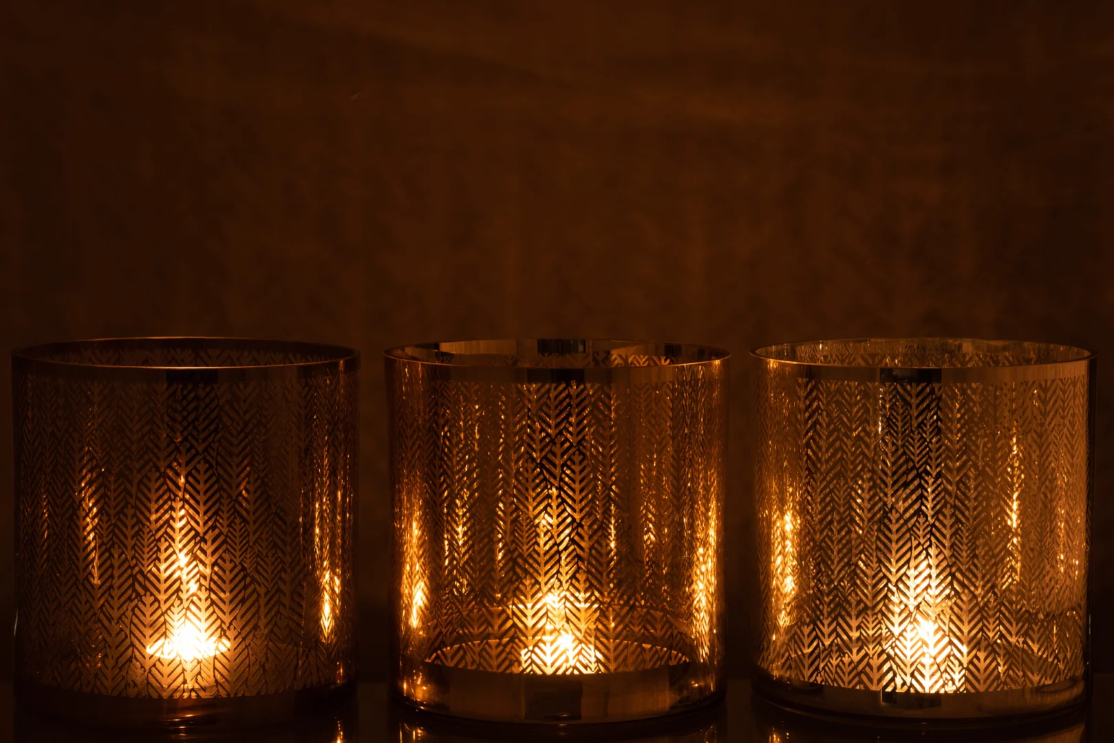 De Kaarsenhouder Glas Graanmotief Zilver Goud Grijs Set 3 heeft drie kaarsen in houders van gestructureerd glas met een korrelmotief dat het licht zachtjes verspreidt en een warme, sfeervolle gloed creëert tegen een donkere achtergrond.