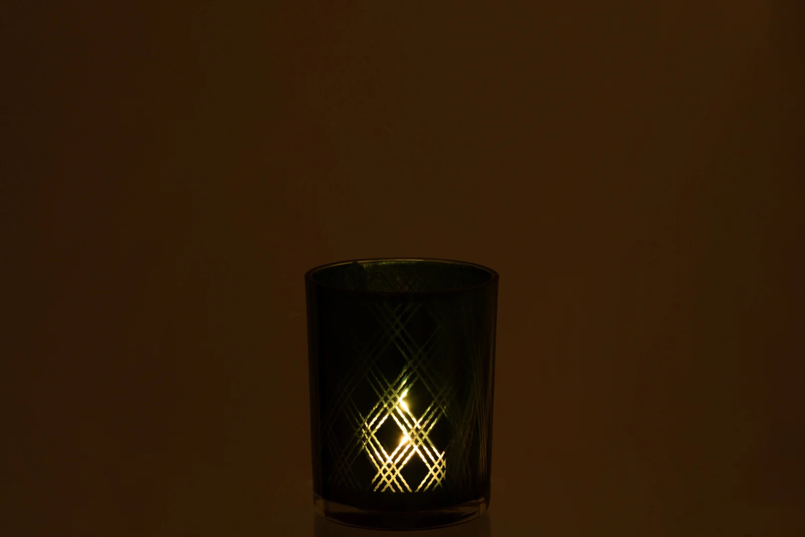 De Theelichthouder Ruit Glas Groen Middel is een donkergroene glazen kandelaar met een brandende kaars erin, die warm licht en kriskraspatronen op het glas werpt, tegen een effen donkere achtergrond.