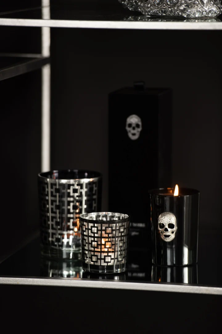 Twee Theelichthouder Vierkant Glas Zilver Zwart Klein kandelaars met geometrische patronen en een derde houder met doodshoofdmotief staan op een glazen plank tegen een donkere achtergrond, met één brandende kaars.