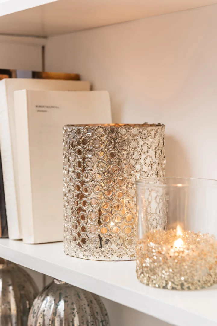 De Kaarsenhouder Orkaan Cirkels Glas Zilver, een metalen glazen kandelaar met ingewikkelde patronen, houdt een brandende kaars op een witte plank naast rechtopstaande boeken.