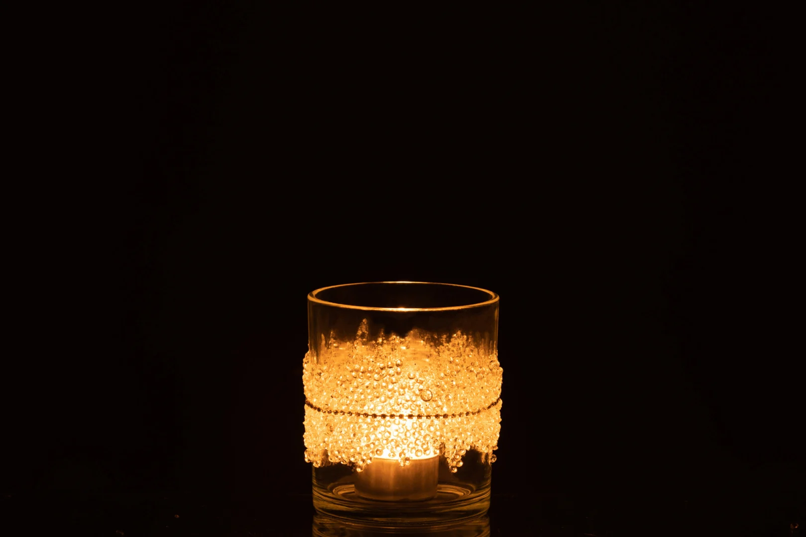 De Theelichthouder Suiker Midden Diamanten Lijn Glas Helder Medium houdt een gloeiende kaars vast. Het heldere glas is versierd met matte, ijzige patronen en diamanten accenten, die afsteken tegen een volledig zwarte achtergrond.