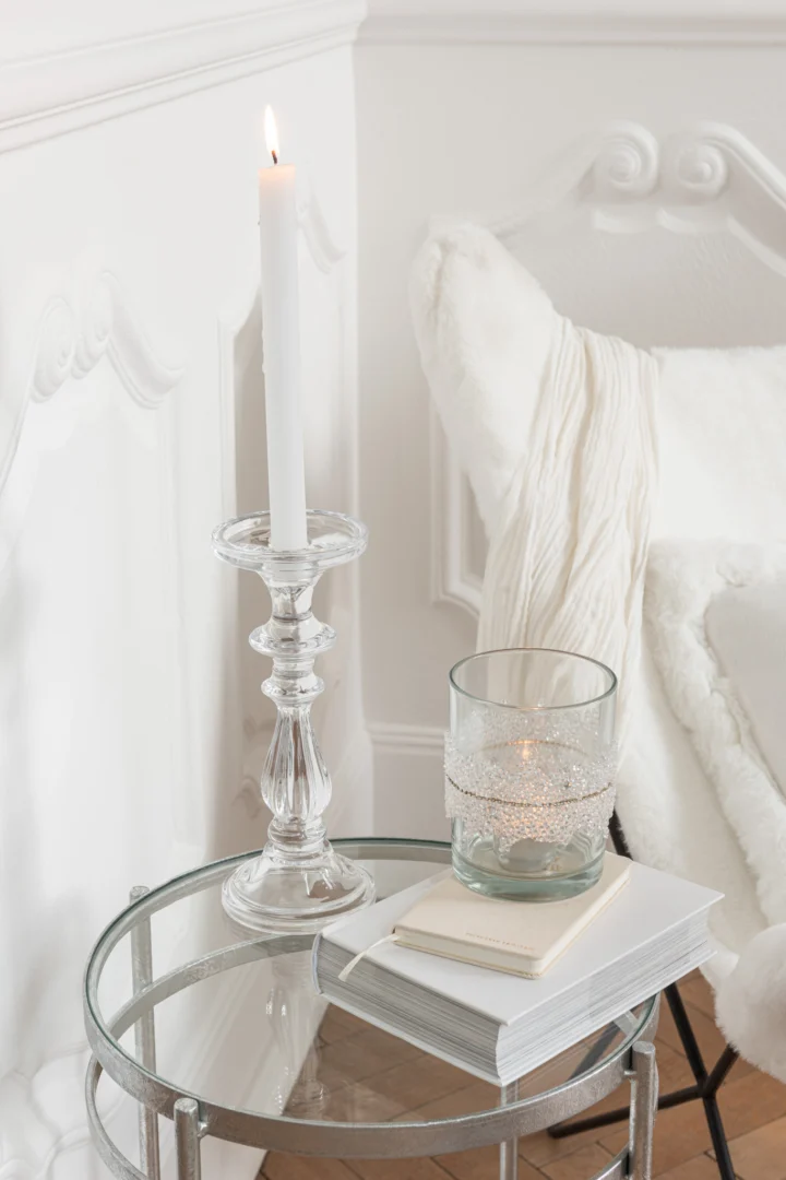 Een brandende witte kaars in Theelichthouder Suiker Midden Diamanten Lijn Glas Helder Medium staat naast een glas met een waxinelichtje en twee gestapelde boeken op een rond glazen bijzettafeltje bij een wit gestoffeerde stoel met een knusse plaid, waardoor een heldere, minimalistische scène ontstaat.