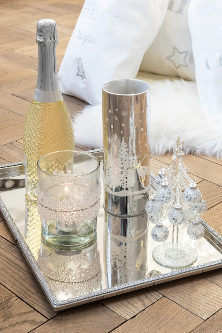 Op een zilveren dienblad staan een fles mousserende wijn, een glas, de Theelichthouder Suiker Midden Diamanten Lijn Glas Helder Medium kandelaar en een glazen kerstboom, op een houten vloer met witte kussens en bont op de achtergrond.