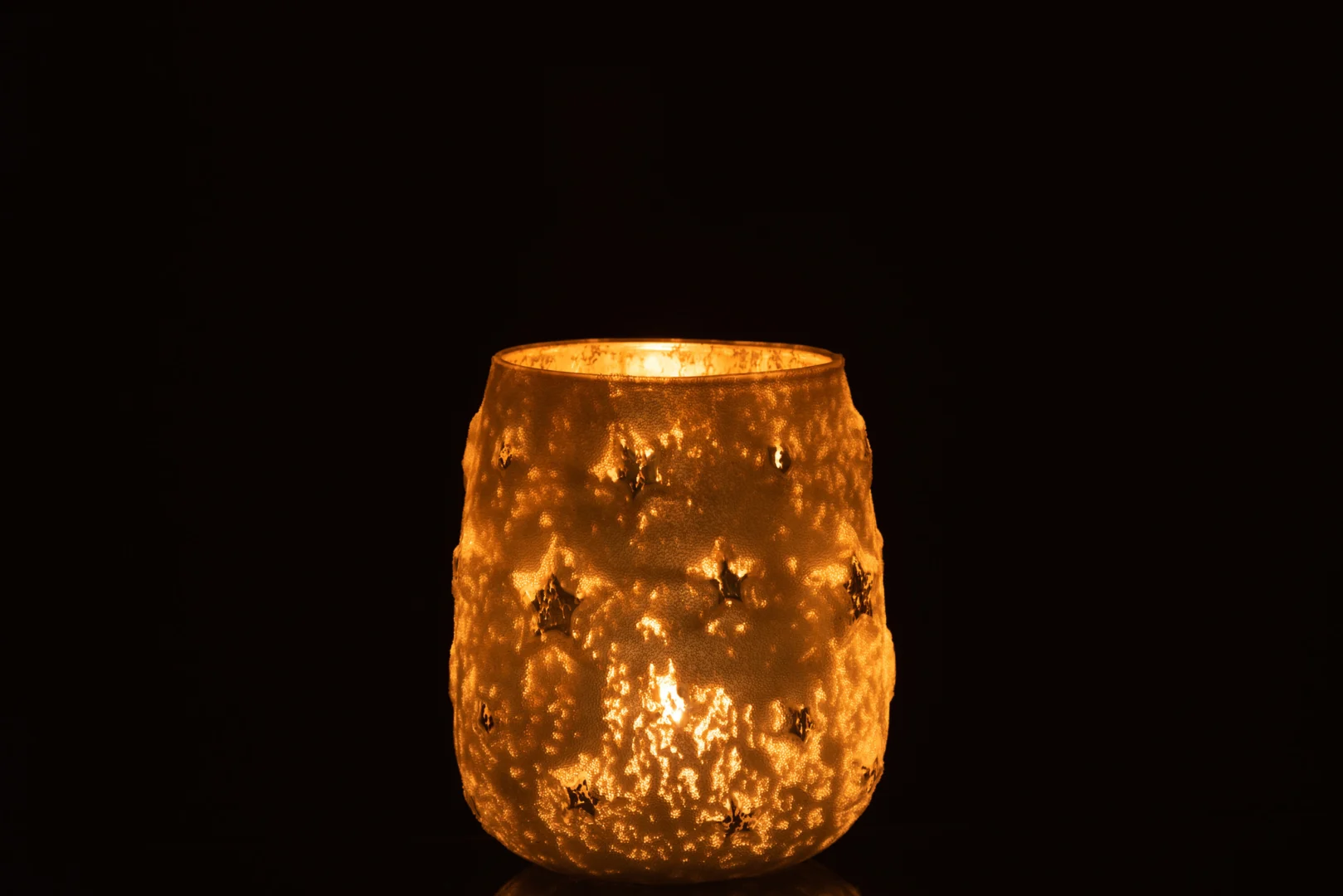 Theelichthouder Ster Glas Antiek Zilver Groot heeft een gestructureerd glazen oppervlak met stervormige uitsparingen, die een warme gouden gloed werpen tegen elke donkere achtergrond.