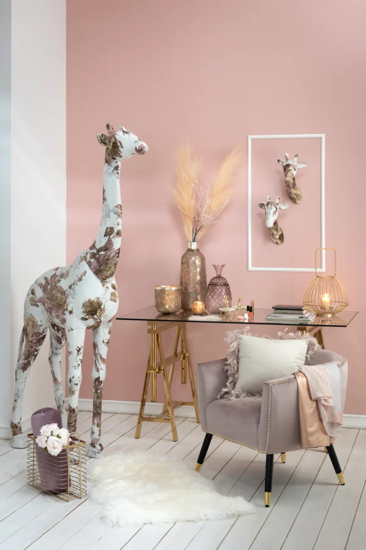 Een stijlvolle kamer met een roze muur is voorzien van Theelichthouder Frosty Glas Oudroze Groot, een giraffesculptuur met bloemmotief, een modern glazen bureau met gouden poten, decoratieve vazen, ingelijste giraffekoppen, een pluche fauteuil en zachte neutraalgetinte accenten.