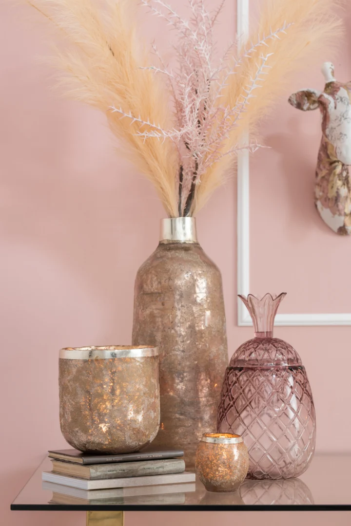 Een glazen tafel toont een stapel boeken, drie metalen kandelaars, de Theelichthouder Frosty Glas Oudroze Groot en een hoge vaas met pampagras, allemaal tegen een roze muur.