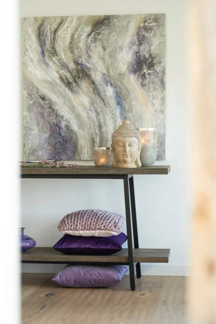 Een decoratieve tafel met daarboven de Theelichthouder Frosty Ronde Glas Zilver Extra groot, een boeddhabeeld en abstracte kunst. Op de plank eronder liggen twee paarse kussens en een glazen vaas voor een rustige, moderne uitstraling.