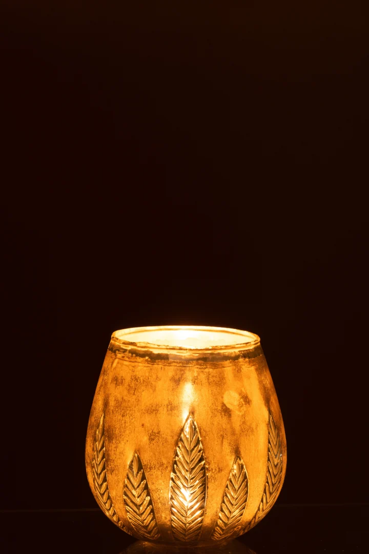 De Theelichthouder Blad Rond Glas Zilver Middel is voorzien van bladmotieven en straalt een warm, gloeiend licht uit dat een opvallend contrast vormt tegen een donkere achtergrond.