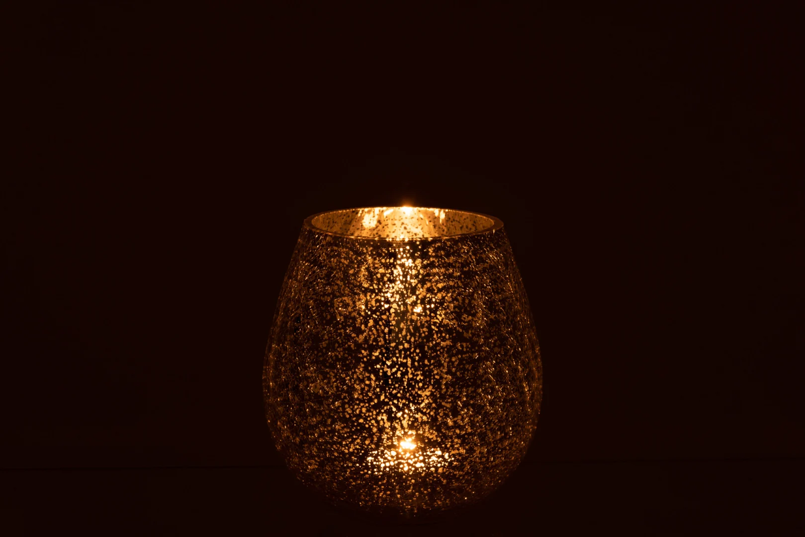 De Kaarsenhouder Orkaan Lichtbal Craquelure Glas Zilver Klein werpt een zachte gouden gloed door het gestructureerde zilveren glas, en creëert zo een warme en uitnodigende sfeer tegen een donkere achtergrond.