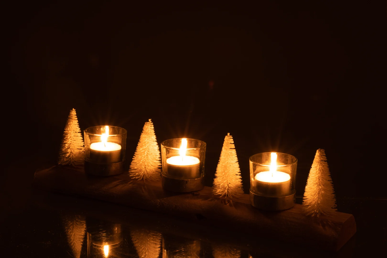 Theelichthouder Kerst Hout Metaal Wit Zilver heeft vier glazen kandelaars op een houten voet, afgewisseld met kleine witte kerstboompjes tegen een donkere achtergrond.