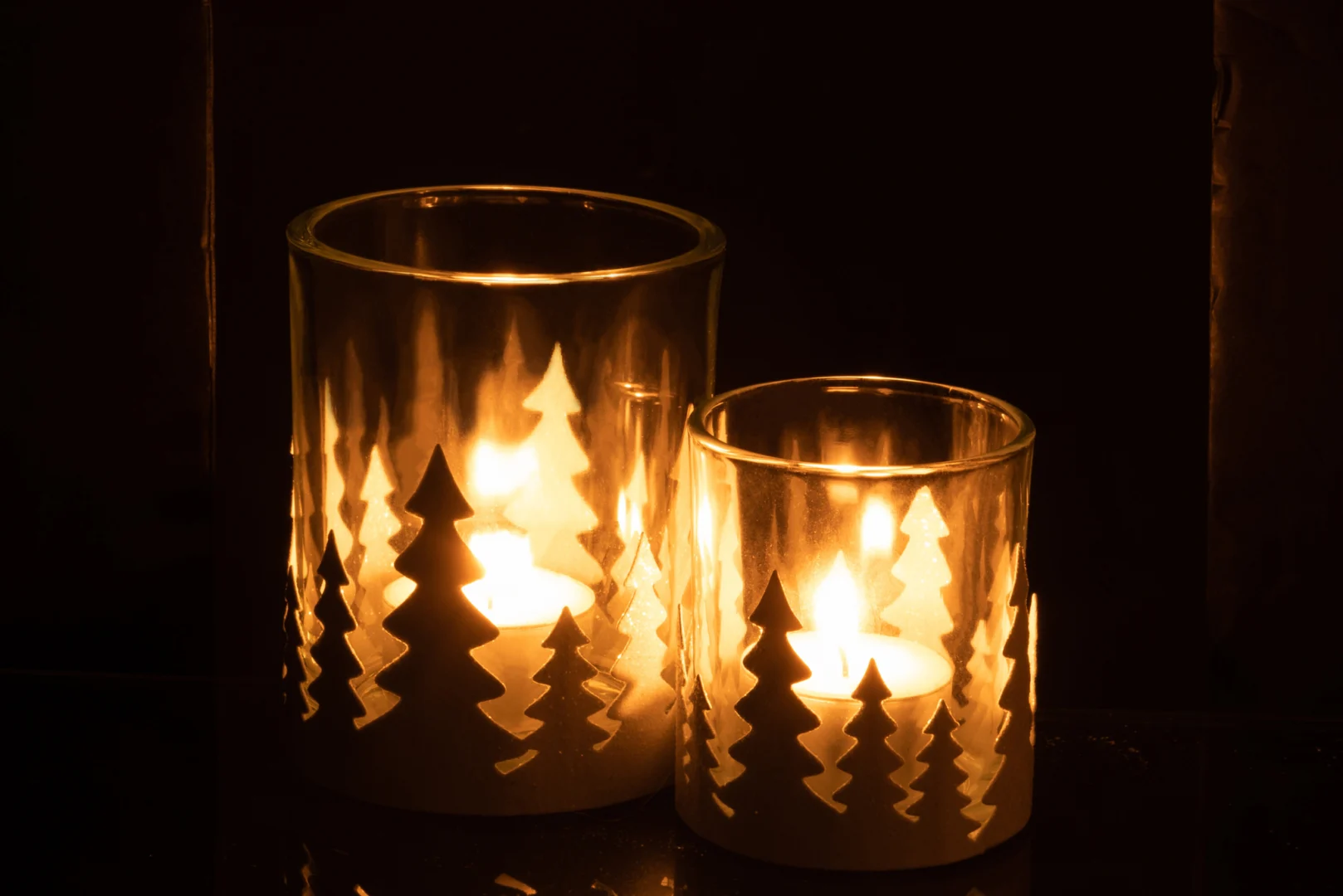 De Theelichthouder Kerstbomen Metaal Glitter Wit Groot heeft twee brandende kaarsen in glazen houders met metallic witte kerstboomuitsnijdingen, die een warme, gloeiende sfeer creëren in het donker.