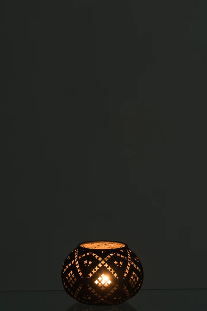 De Theelichthouder Bal Oosters Glas Bruin Antiek zit onderaan de afbeelding, gloeit zachtjes met een kaars erin en werpt ingewikkelde lichtpatronen tegen een donkere achtergrond.
