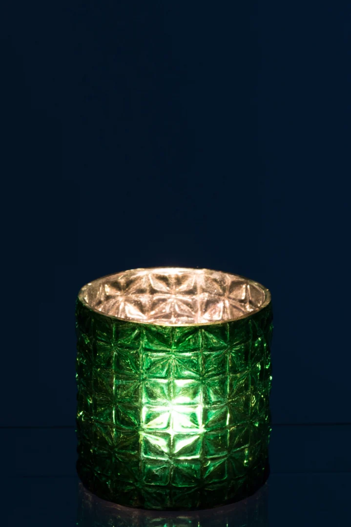 De Kaarsenhouder Orkaan Patroon Glas Groen Klein gloeit zacht van binnenuit, het groen getextureerde glas werpt zacht licht tegen een donkerblauwe achtergrond.