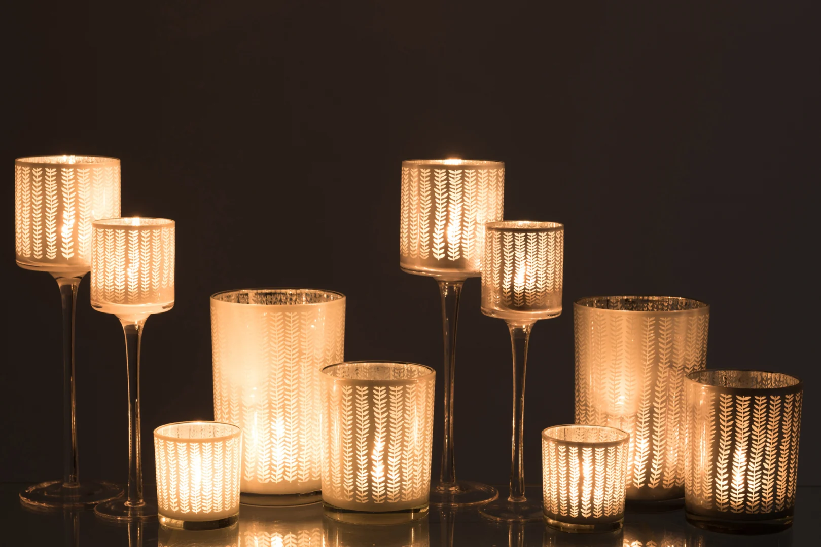 De Kaarsenhouder Orkaan Sarah Glas Wit Blauw Groot Set 2 bestaat uit decoratieve glazen kandelaars van verschillende grootte, elk warm oplichtend met kaarsen erin, prachtig uitgestald tegen een donkere achtergrond.