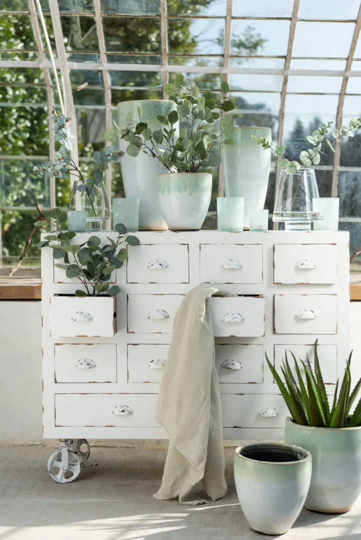 Een set van twee grote ijsblauwe glazen waxinelichthouders (Theelichthouder Glas IJsblauw Groen Groot Set 2) staat op een vintage witte ladekast, naast pastelkleurige potplanten en een aloë. Zonlicht valt naar binnen door brede glazen ramen.