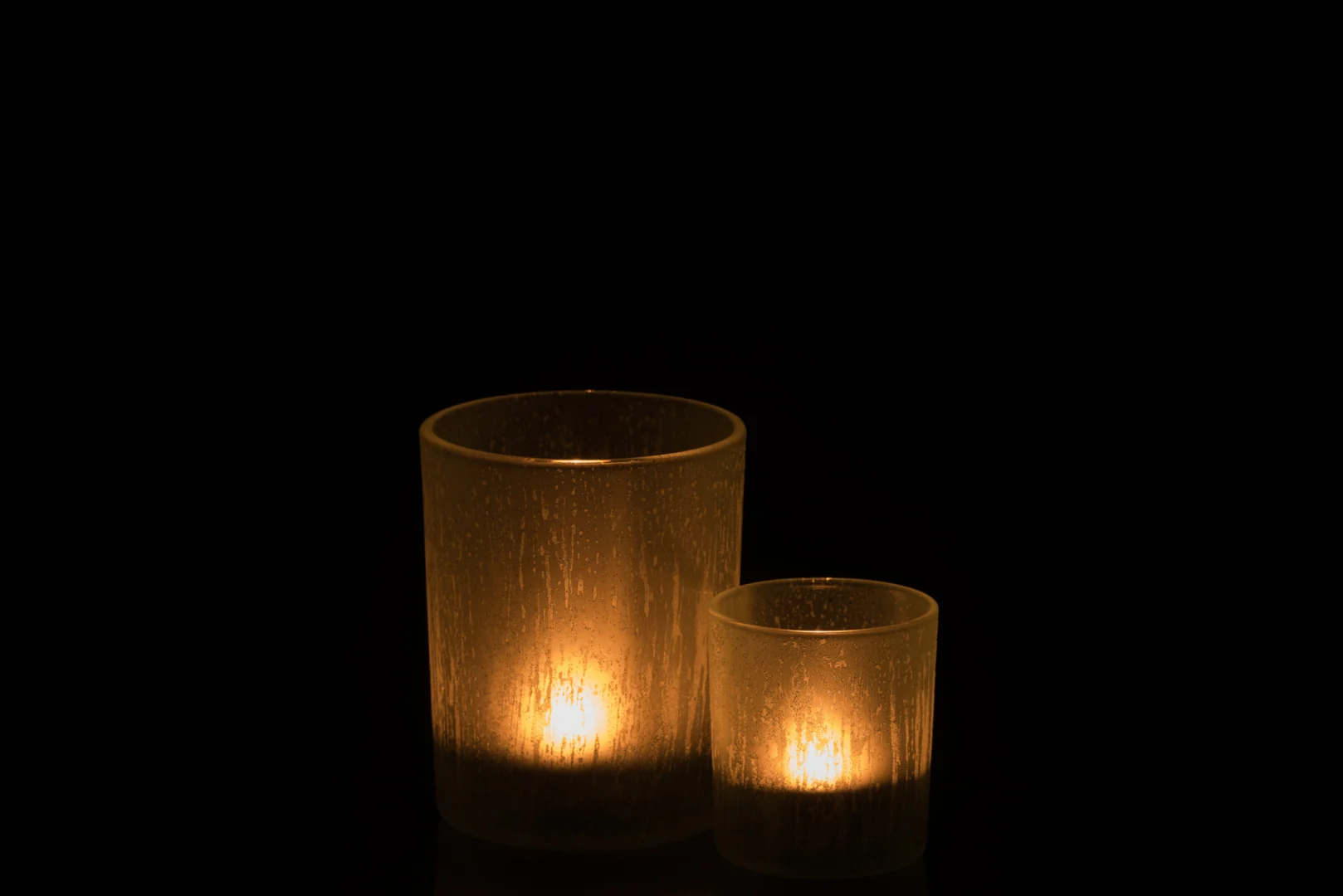 De Theelichthouder Glas IJsblauw Groen Groot Set 2 heeft twee gloeiende kaarsen - een hoge, een korte - in ijsblauwe en groene glazen houders, die een warme sfeer creëren tegen een zwarte achtergrond.