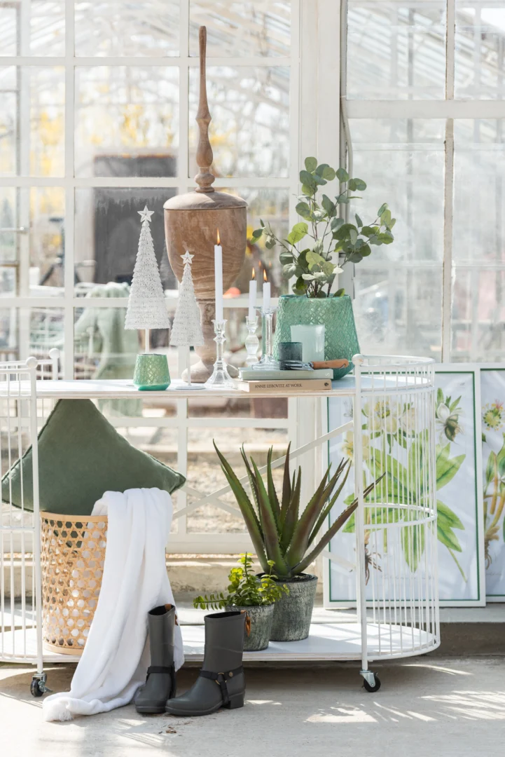 Een lichte serre heeft een witte metalen kar met groene potplanten, boeken, decoratieve bomen en Theelichthouder Glas IJsblauw Groen Groot Set 2 kaarsen; hieronder staan regenlaarzen, een mand met kussen en deken, plus botanische prints bij het raam.