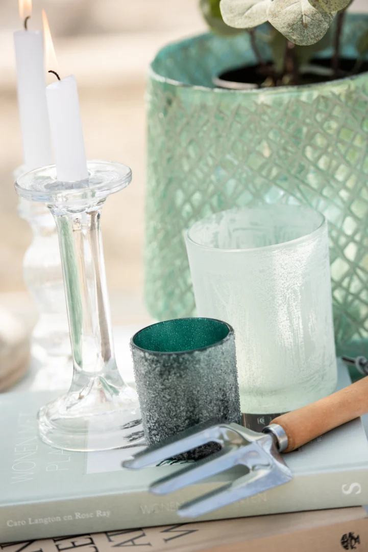 Een tuinvork rust op boeken naast Theelichthouder Glas IJsblauw Groen Groot Set 2, een groene votief, een mat glas en een groene plantenpot met structuur en een witte kaars in een glazen kandelaar.
