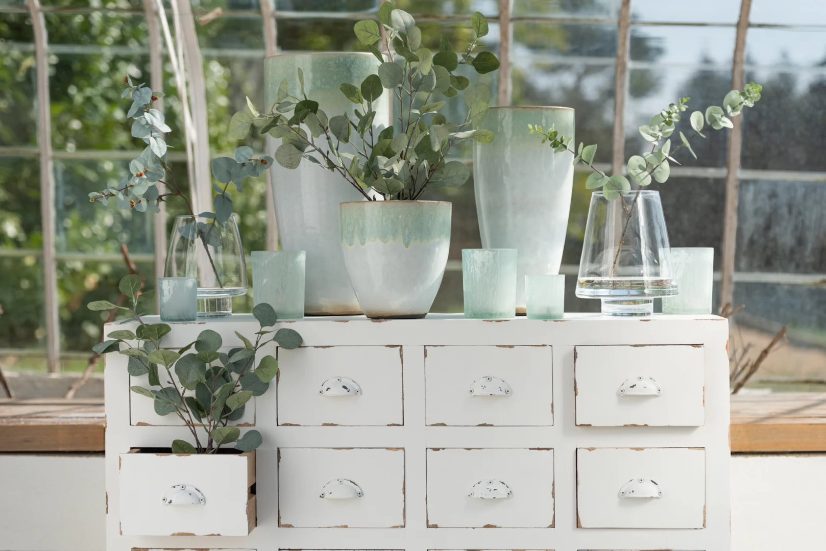 De Theelichthouder Glas IJsblauw Groen Groot Set 2, uitgestald op een vintage witte kast met verweerde lades, is een aanvulling op eucalyptusstengels, allemaal voor grote ramen met weelderig groen buiten.