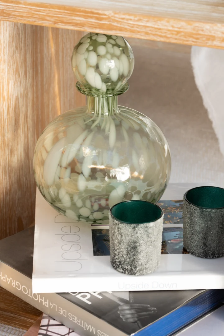 De Theelichthouder Parelglas Zwart Mat Klein, met een rond zwart mat gespikkeld ontwerp, rust op gestapelde boeken naast twee kleine bekers met structuur en een donkergroene binnenkant op een houten oppervlak.