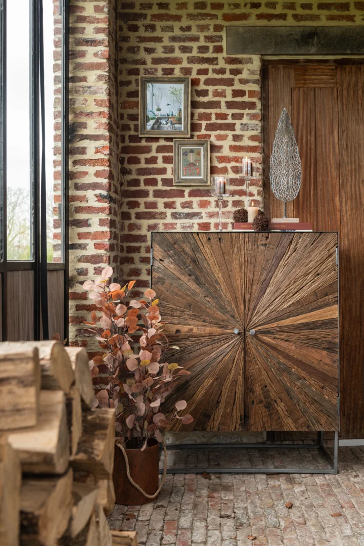 In een rustieke kamer met bakstenen muren staat een houten kast, gestapeld haardhout, ingelijste kunst erboven en de Kaarsenhouder Op Voet Cilinder Glas Transparant Klein naast een potplant met koperen bladeren.