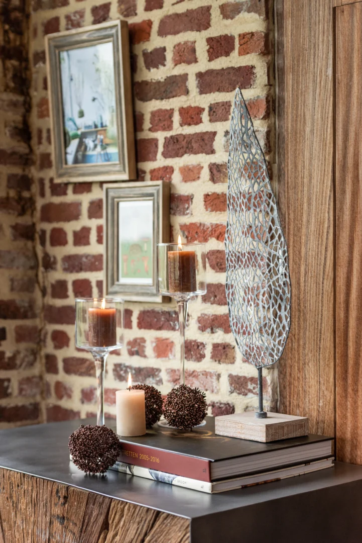 Een moderne decoropstelling met de Kaarsenhouder Op Voet Cilinder Glas Transparant Medium, decoratieve bollen, een metalen bladsculptuur en gestapelde boeken op een metalen oppervlak, tegen een bakstenen muur met ingelijste kunst.