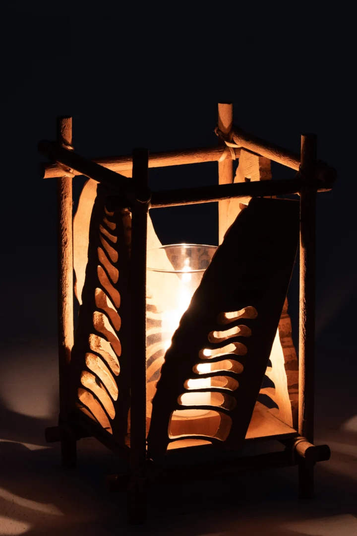 De Kaarsenhouder Bamboe Glas Natuurlijk Groot heeft een kaars in een glazen houder in een frame van natuurlijk bamboe met uitgesneden bladeren, die warme, ingewikkelde schaduwen werpt in het donker.