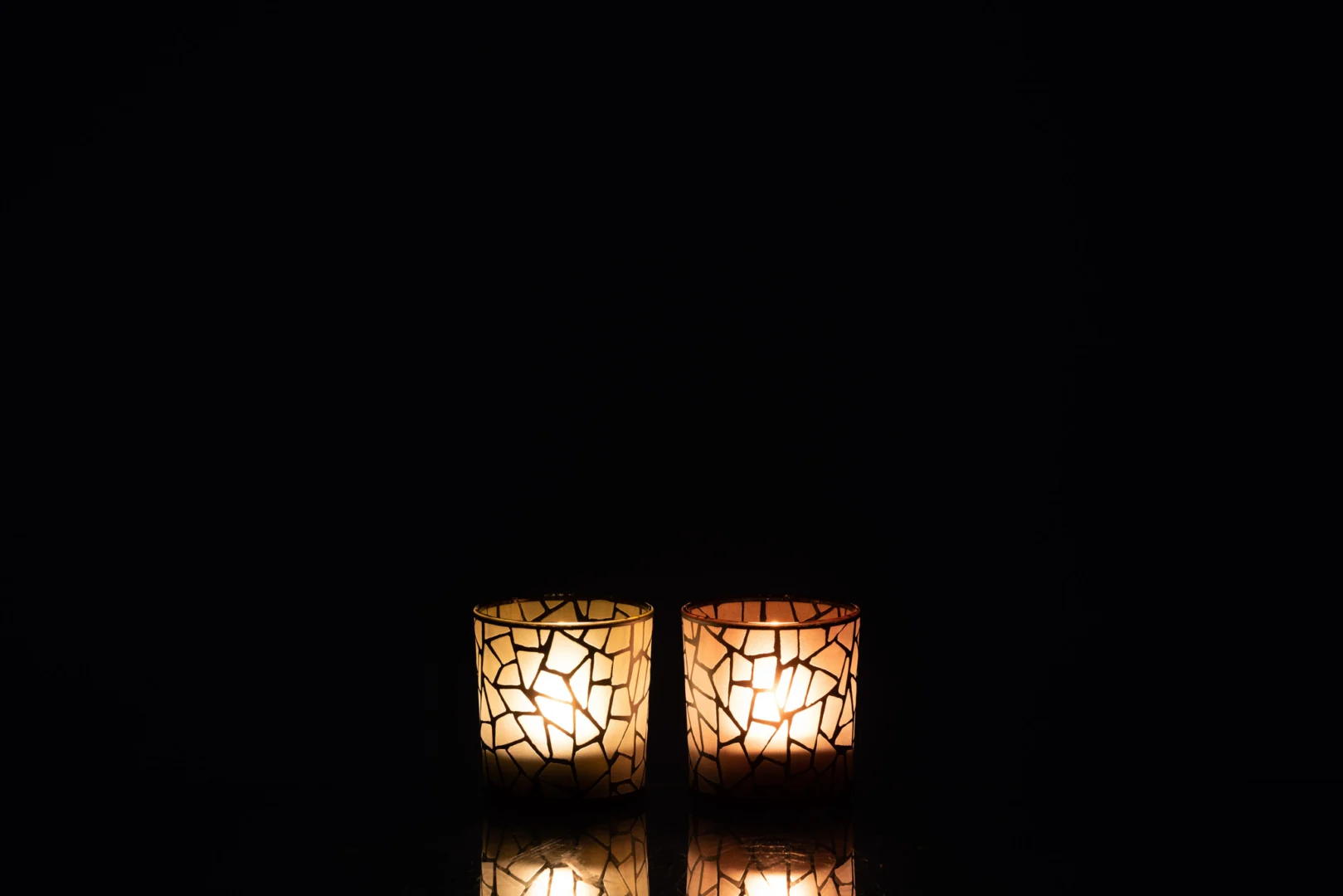 Theelichthouder Mozaïek Glas Oker Bruin Klein Set van 2 staat naast elkaar tegen een zwarte achtergrond, waarbij hun warme gloed en zachte reflecties zichtbaar zijn op het oppervlak eronder.