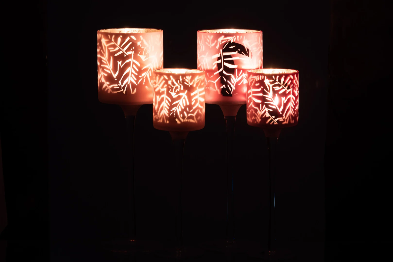 Set van 2 Theelichthouder Op Voet Toekan Glas Roze Groot kandelaars stralen warm met hun bladachtige uitgesneden patronen en werpen prachtig decoratief licht en schaduw tegen een donkere achtergrond.