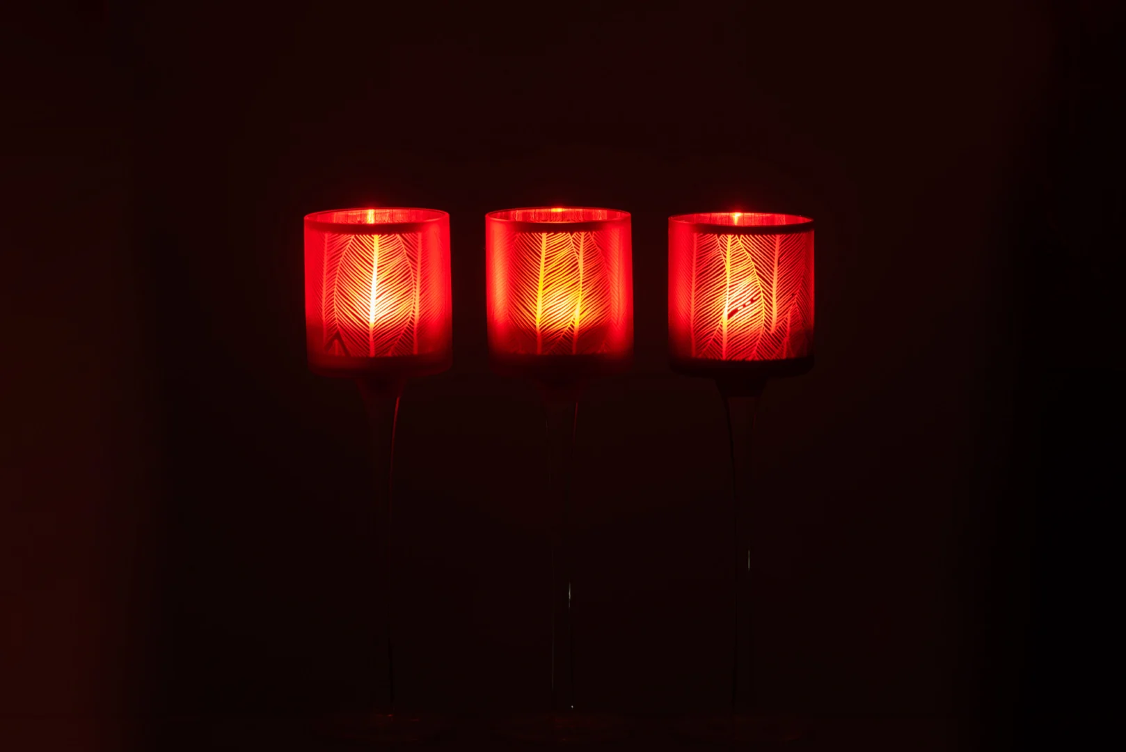 Theelichthouder Op Voet Bladeren Glas Rood Set 3 heeft drie rode glazen kandelaars op een rij die warm oplichten en decoratieve patronen vormen tegen een donkere achtergrond.