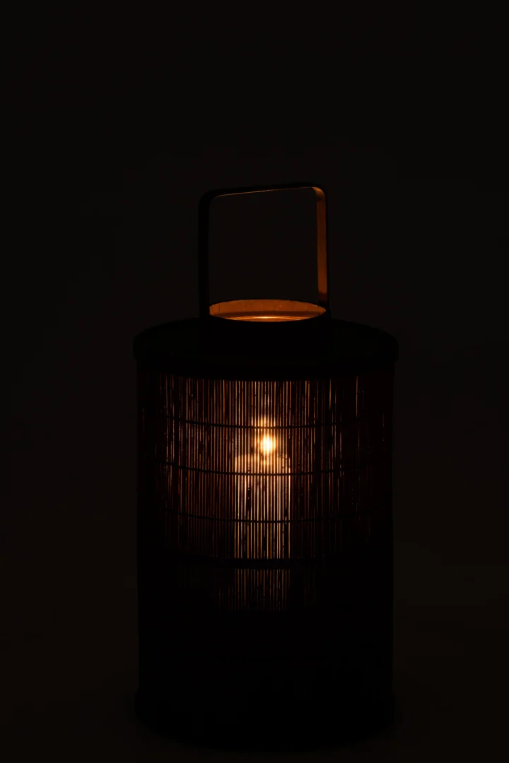 Een brandende kaars gloeit in de Kaarsenhouder Lantaarn Cilinder Bamboe Bruin Groot, een ronde, gevlochten lantaarn met een handvat, tegen een donkere achtergrond.