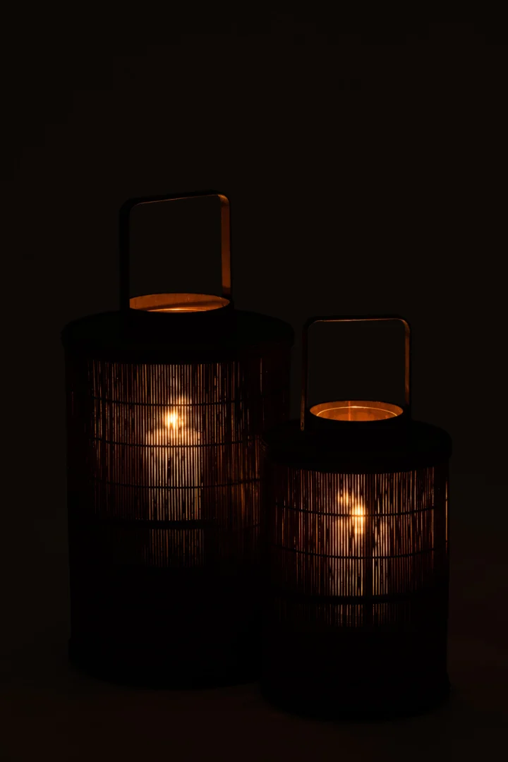 De Kaarsenhouder Lantaarn Cilinder Bamboe Bruin Groot gloeit warm in het donker, met verticale bamboe latten, een handvat bovenop en een zichtbare kaars binnenin die zacht, sfeervol licht werpt door de latten.