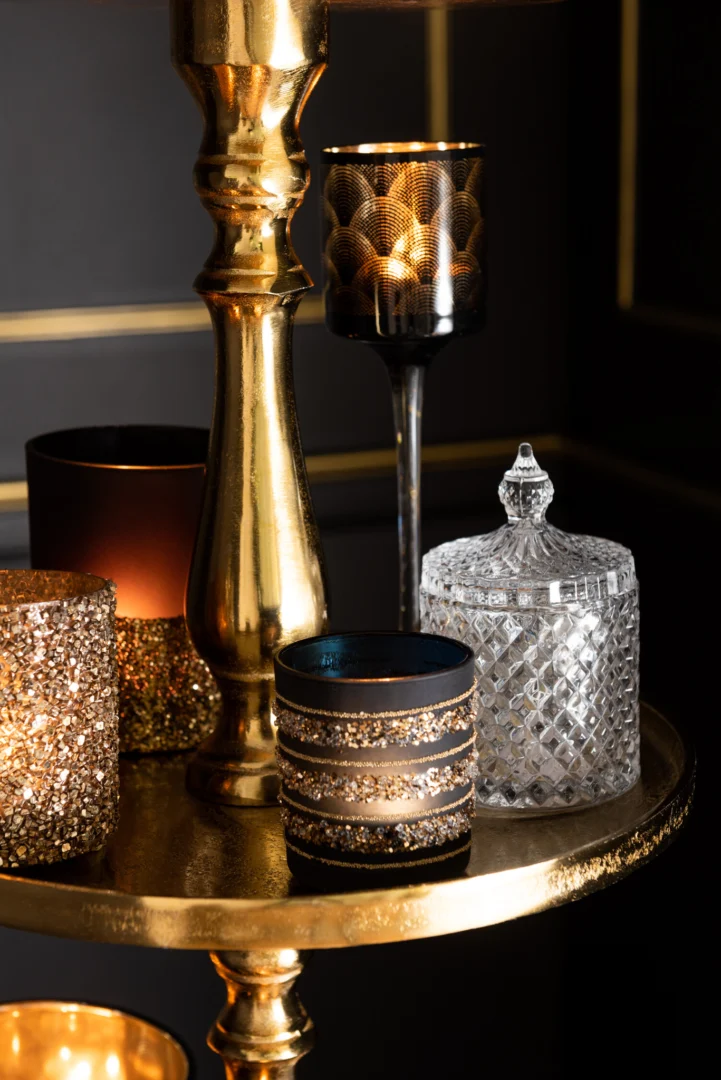 Op een gouden dienblad staat de Theelichthouder Op Voet Gatsby Glas Zwart Goud Klein Set 2, gecombineerd met een glazen pot en kaarsen in glitterende houders, die een warme en elegante sfeer creëren.
