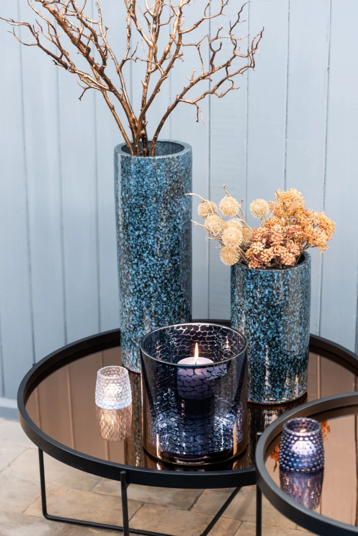 Op een ronde metalen tafel staat de Kaarsenhouder Orkaan Slang Glas Blauw Medium met een brandende blauwe kaars, blauwe glazen vazen gevuld met gedroogde takken en bloemen en twee kleine waxinelichthouders, allemaal tegen een lichtblauwe houten muur.