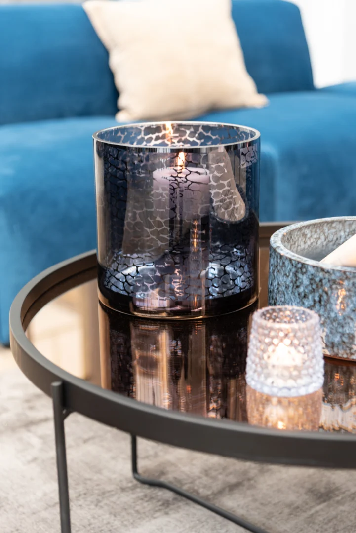 Op een moderne salontafel staat de Kaarsenhouder Orkaan Slang Glas Blauw Medium met een brandende kaars, naast twee kleinere glazen houders met structuur. Een blauwe bank met een licht kussen staat op de achtergrond en creëert een gezellige en stijlvolle sfeer.