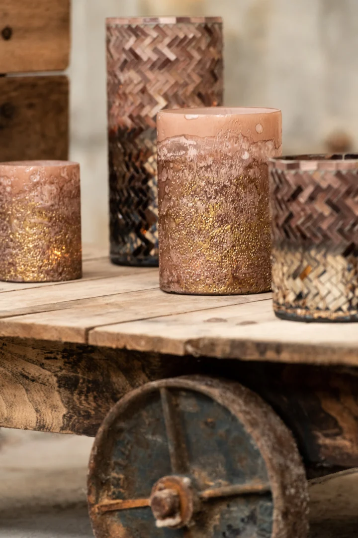 De Kaarsenhouder Orkaan Lijn Glas Roze Goud Klein, een set van vier decoratieve glazen kandelaars met roze en gouden accenten, staat op een rustieke houten kar met een metalen wiel tegen een neutrale achtergrond.