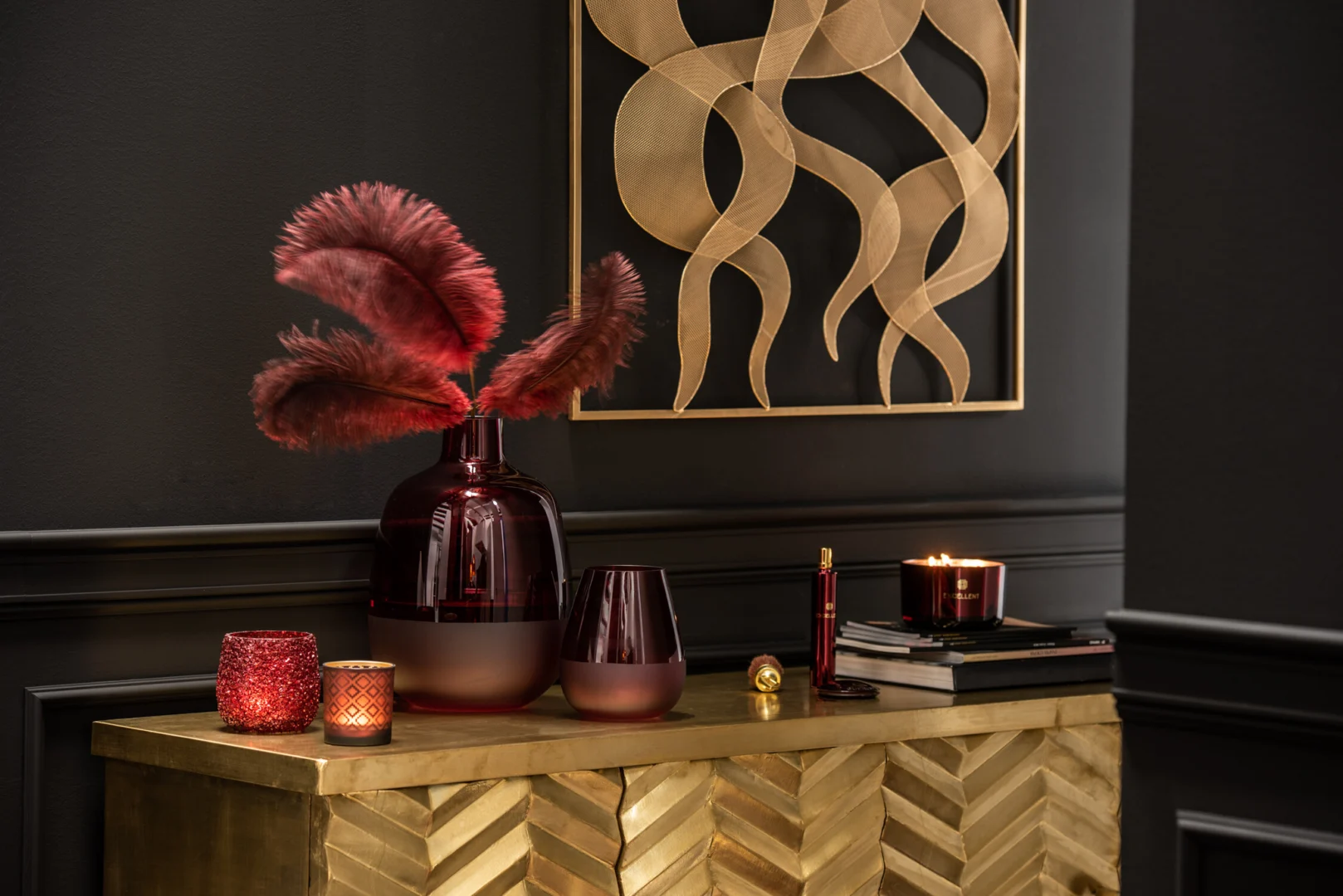 Een gouden kist met visgraatmotief bevat Theelichthouder Glitter Glas Rood, een wijnglas, een grote donkere vaas met rode veren en boeken; gouden abstracte muurkunst siert de donkere muur erachter.